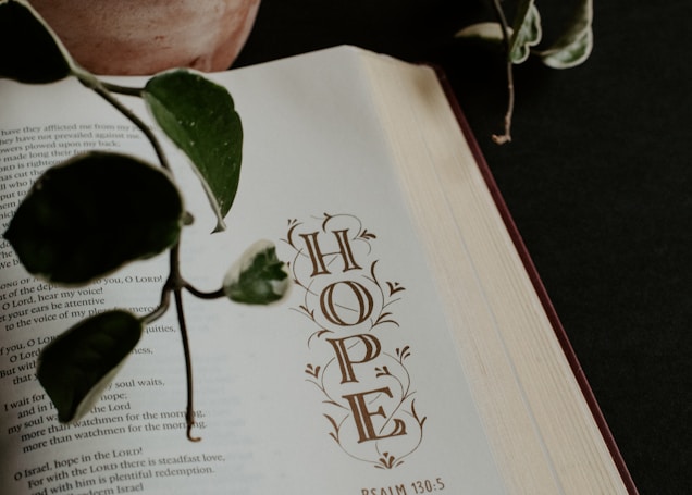 An open book showing a page with the word 'HOPE' and a reference to Psalm 130:5. A potted plant with green leaves extends its branches over the book.